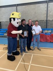 Image: A life-size mascot of a miner stands with three other men, two of them holding plaques recognizing their awards for Honorary King Miner and Honorary Driller for Nickel Days 2022 in Thompson, Man.