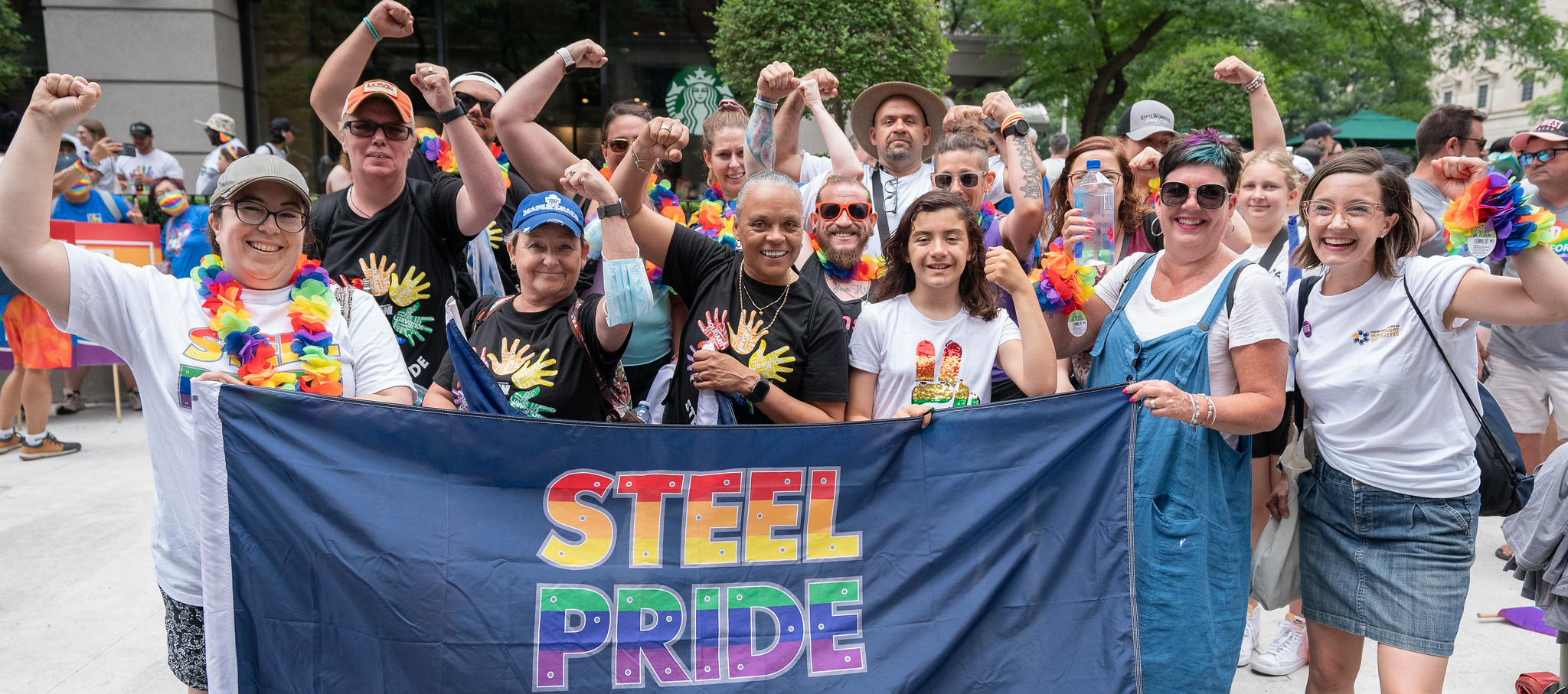 A group of people standing outdoors, with their fists up. There is a flag they are holding up that says "Steel Pride"