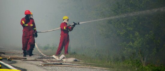 Three people in red jumpsuits with reflective yellow tape and wearing yellow hardhats are in the foreground. The person on the right is spraying water from a hose to the right. In the background is heavy smoke obliterating any details in the landscape. Photo: Canadian Red Cross