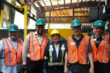 Five people standing inside an industrial building. They are all wearing safety vests and hard hats.